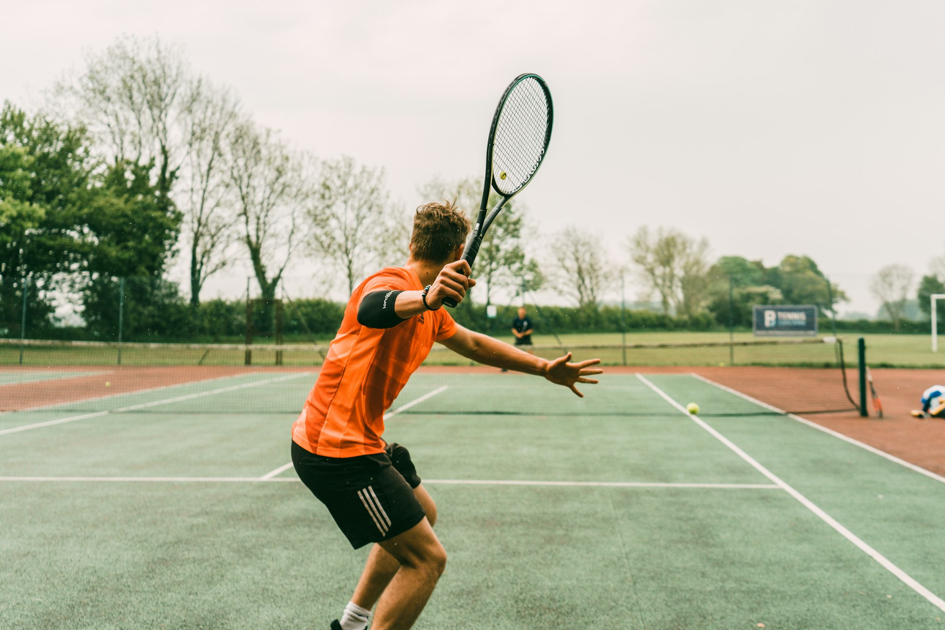 A tennis player stretching for a forehand on an outdoor court lined with trees
