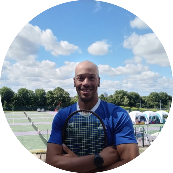 Coach Falco smiling, arms folded around his tennis racket, in front of outdoor tennis courts on a sunny day